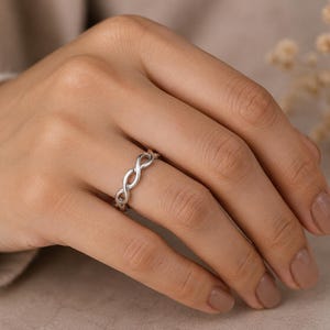 May include: A polished silver ring featuring a continuous infinity symbol pattern that wraps around the entire band. The metal has a smooth, reflective finish, and the design consists of interconnected loops forming a repeating figure-eight motif.