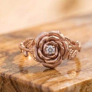 May include: A rose-shaped ring crafted from rose-colored metal, featuring a textured, layered petal design that mimics a blooming flower. A single, small, clear round-cut gemstone is set securely in the center of the rose, with delicate leaf details.