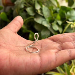 Puede incluir: Un anillo de metal de color plateado con un diseño de alambre continuo que forma una base circular y se eleva en un solo bucle vertical. El anillo está construido con una sola pieza de alambre pulido, creando una forma simple y abierta.