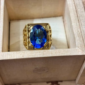 Pode incluir: Um anel de tom dourado com uma grande pedra preciosa azul-escura de corte oval no centro. A banda tem uma face retangular larga com um padrão geométrico vazado nas laterais, exibindo um acabamento metálico e uma pedra azul facetada.