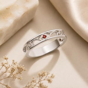 May include: A silver-toned ring featuring an intricate openwork scroll pattern along the band. A single small, round, deep red gemstone is set securely within the centre of the metal filigree design, creating a contrast against the polished silver.