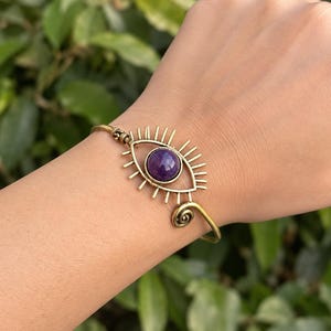 May include: A brass-toned metal cuff bracelet featuring an eye-shaped design with a central round purple gemstone. The metal frame radiates outward like eyelashes, and the ends of the open cuff are finished with small, coiled spiral details.