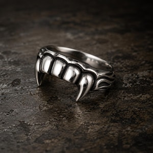 May include: A silver-toned metal ring featuring a sculpted design of a row of teeth with two prominent fangs extending downward. The surface of the ring has a polished finish on the teeth and a darkened, oxidized texture on the band sides.