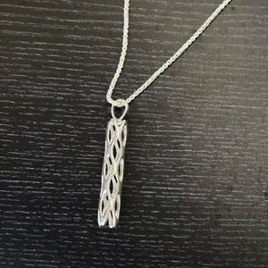 May include: A silver-toned necklace featuring a slender, cylindrical pendant with an intricate, openwork lattice design. The pendant hangs from a delicate, thin silver-toned chain, showcasing a repeating, interconnected oval pattern throughout its length.