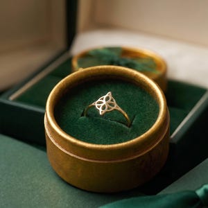 May include: A gold ring featuring a delicate, openwork four-petaled floral motif on the band. The ring is presented inside a round, gold-colored container with a deep green velvet interior lining, designed to hold jewelry securely in place.