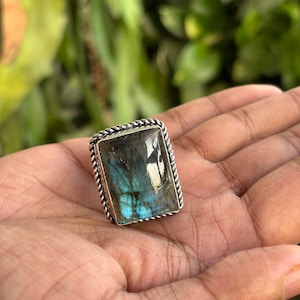 Puede incluir: Un anillo de labradorita rectangular con una piedra pulida e iridiscente en tonos azules y grises. La piedra está engastada en un marco de metal plateado con un borde decorativo de estilo cuerda trenzada alrededor del contorno exterior.
