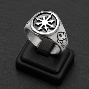 Op de afbeelding: Een zilverkleurige metalen ring met een cirkelvormig voorvlak met een verhoogd achtpuntig stersymbool tegen een donkere, verzonken achtergrond. De ringband vertoont gedetailleerde zwarte gegraveerde lijntekeningen op de zijkanten.