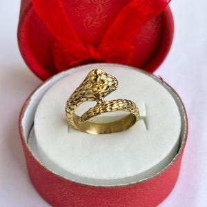 Peut inclure: Une bague en métal doré en forme de serpent enroulé avec un motif d'écailles texturé sur sa surface. La bague est présentée à l'intérieur d'une boîte cadeau ronde rouge vif avec un nœud en ruban rouge assorti sur le couvercle, sur un insert blanc.