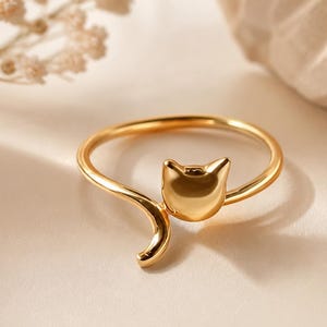 May include: A gold-toned ring featuring a smooth, stylised cat head design with pointed ears and a curved tail that wraps around the finger. The metal surface is polished and reflective, creating a sleek, continuous band shape.