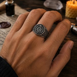 Op de afbeelding: Een zilverkleurige metalen ring met een cirkelvormig voorvlak met een gegraveerde zespuntige ster, omsloten door een slangenmotief. De band vertoont aan de zijkanten ingewikkelde knoop patronen met een donkere, geoxideerde textuur.