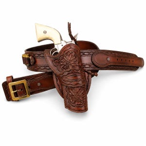 May include: A brown leather holster and belt set featuring intricate floral tooling and a brass buckle. A revolver with a cream-coloured grip is secured within the holster. The belt includes loops holding several metallic cartridges on the side.