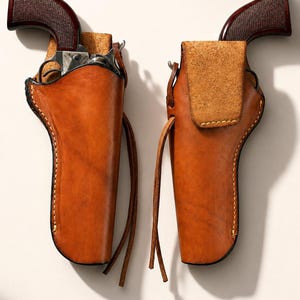 Può includere: Due fondine in pelle color cuoio abbinate, ciascuna contenente un revolver con impugnatura testurizzata marrone scuro. Le fondine presentano cuciture visibili lungo i bordi, una chiusura a patta sul lato destro e lunghi lacci in pelle.