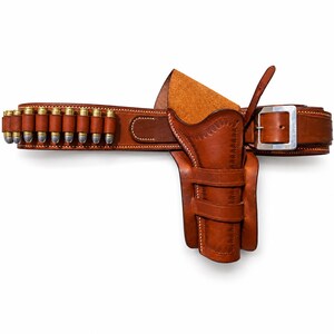 Op de afbeelding: Een bruin lederen pistoolholster bevestigd aan een bijpassende riem met een reeks lussen voor metalen patronen. De holster is voorzien van decoratieve stiksels langs de randen en een zilverkleurige metalen gesp op de riem voor verstelling.