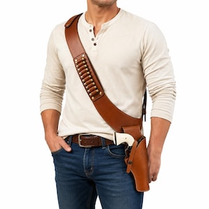Op de afbeelding: Een lichtbruine lederen schouderholster met een brede schouderband en geïntegreerde lussen voor twaalf patronen met messing hulzen. De holster is aan de heup bevestigd en bevat een revolver met een crèmekleurige greep en een metalen loop.