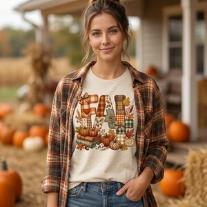 Op de afbeelding: Een crèmekleurig t-shirt met een afbeelding van het woord FALL in een patchworkpatroon met pompoenen, boeken en herfstbladeren. Het t-shirt wordt gedragen onder een opengeknoopt flanellen overhemd met bruin, oranje en zwart ruitpatroon.