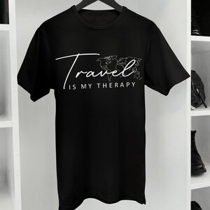 May include: A black short-sleeved t-shirt featuring a white graphic print of a world map outline and the text "Travel IS MY THERAPY" in a combination of script and sans-serif fonts. The garment has a crew neckline and is displayed on a hanger.