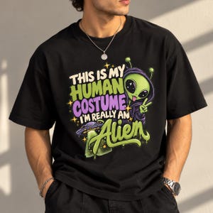 Pode incluir: Uma t-shirt preta com o gráfico de um alienígena verde vestindo um capuz roxo e fazendo um sinal de paz. O texto diz: This is my human costume I'm really an alien. Um pequeno disco voador é visível ao lado do personagem.