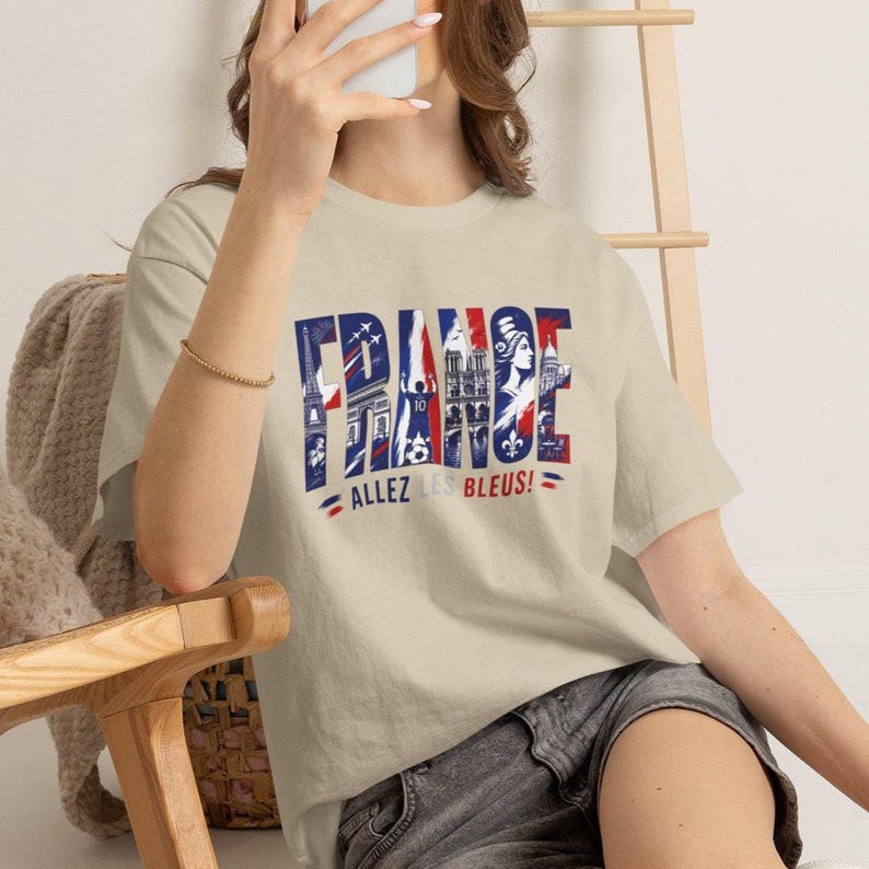 Puede incluir: Una camiseta beige de manga corta con un estampado gr&aacute;fico de la palabra FRANCE en azul, blanco y rojo. Las letras contienen ilustraciones de la Torre Eiffel, un jugador de f&uacute;tbol y Notre Dame. Debajo del texto est&aacute; la frase ALLEZ LES BLEUS!