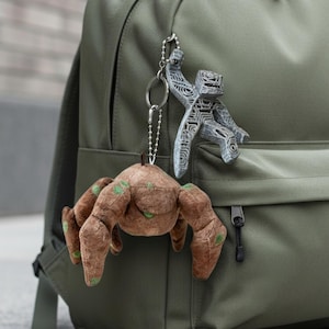 Puede incluir: Mochila verde oliva con un bolsillo frontal y cremalleras negras. Dos llaveros cuelgan de la cremallera: una araña de peluche marrón con detalles verdes y una figura gris estilizada.