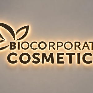 以下が含まれることがあります： 暗い立体的な文字で「BIOCORPORATE COSMETICS」と書かれた壁掛け看板。テキストの左側には、4枚の花びらを持つ花のアイコンがデザインされています。看板全体がバックライトで照らされ、壁面に温かみのある柔らかな光を放っています。