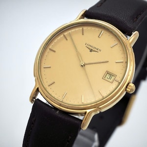 May include: A gold-tone wristwatch with a round champagne-coloured dial, thin gold hour markers, and gold hands. The watch features the Longines logo below the twelve o'clock position, a date window at three o'clock, and a dark brown leather strap.