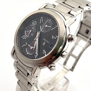 May include: A silver-toned stainless steel Seiko Lukia chronograph watch with a black dial, featuring Arabic numeral hour markers, three sub-dials, a date window, and a metallic link bracelet with a deployment clasp, presented on a white stand.