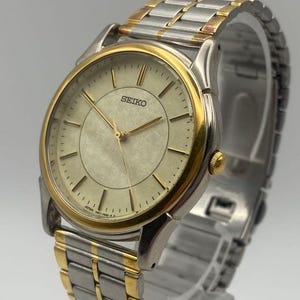 May include: A Seiko wristwatch featuring a round, textured cream-colored dial with gold-tone hour markers and hands. The watch has a two-tone stainless steel link bracelet with silver and gold-colored segments and a gold-tone bezel.