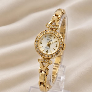 May include: A gold-tone analog wristwatch featuring a round white dial with the text Diana and Orient, gold-tone hands, and Arabic numerals at 3, 6, 9, and 12. The bezel and lugs are embellished with small clear stones, attached to a gold-tone link bracelet.