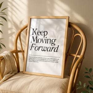 Puede incluir: Una lámina rectangular en un marco de madera clara con el texto negro Keep Moving Forward en una fuente serif, seguido de un párrafo de texto más pequeño que comienza con You may not notice how far you have come while you are still.