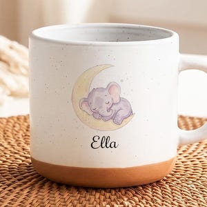 Puede incluir: Una taza de cerámica blanca con acabado moteado y base de color terracota. El frente presenta una ilustración en acuarela de un pequeño elefante morado durmiendo sobre una luna creciente amarilla, con el nombre Ella impreso en negro.