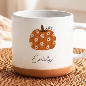 May include: A white ceramic mug with a speckled finish and a terracotta-coloured base. The front features an orange pumpkin illustration decorated with small white daisy flowers and the name Emily written in black cursive script below the pumpkin.