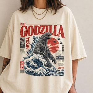 May include: An off-white t-shirt featuring a graphic print of Godzilla emerging from blue ocean waves. The design includes red Japanese text, the word GODZILLA in large red letters, a red sun, helicopters, and small inset images of the creature.
