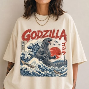 May include: An off-white short-sleeve t-shirt featuring a graphic print of Godzilla emerging from a large blue wave. The design includes the red text "GODZILLA", a red sun, a mountain, helicopters, and Japanese characters. The shirt has a crew neckline.