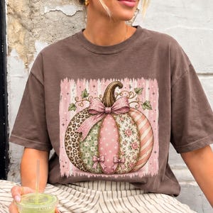 May include: A brown short-sleeve t-shirt featuring a large graphic of a pumpkin with patchwork patterns in pink, green, and leopard print, accented with pink bows. The pumpkin is set against a vertical pink and white striped background.