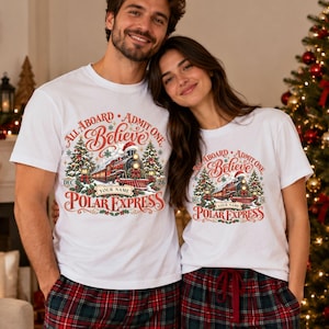 May include: White short-sleeve t-shirts featuring a graphic of a steam train with a Santa hat, Christmas trees, and the text: All Aboard Admit One Believe Polar Express Dec 26 Your Name. The design uses red, green, and gold colors.