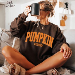 May include: A dark chocolate brown long-sleeve sweatshirt featuring the orange text PUMPKIN SEASON in a distressed, arched font. The garment has a crew neckline and long sleeves, and the text is centred on the chest of the sweatshirt.