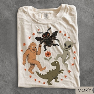 May include: An ivory t-shirt featuring a graphic print of a brown Bigfoot, a black moth-like creature, a grey alien, and a green dinosaur arranged in a circle with orange stars and a central orange flower. The inner neck label reads Comfort Colors.