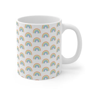 Könnte beinhalten: Weiße Keramik-Tasse mit einem sich wiederholenden Muster aus bunten Regenbogen. Die Regenbogen sind in den Farbtönen Rot, Orange, Gelb, Grün, Blau und Lila gehalten. Die Tasse hat einen gebogenen Henkel und eine glatte, glänzende Oberfläche.