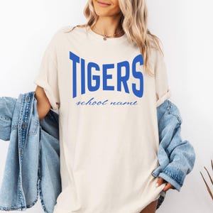 May include: A cream-colored short-sleeve t-shirt featuring the word TIGERS in large, bold, blue capital letters across the chest, with the words school name printed in a smaller, blue, cursive script font directly underneath the main text.