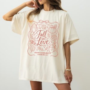 May include: An off-white short-sleeve t-shirt featuring a red line-art graphic of autumn elements, including a pumpkin, mugs, and leaves. The text reads Fall in Love, Sabina's Bachelorette, and Jackson Hole, WY, enclosed in a decorative ribbon frame.