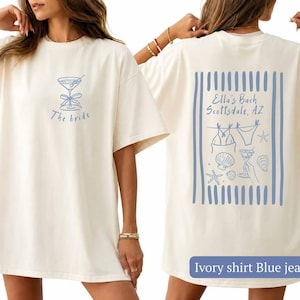 May include: An ivory t-shirt featuring a blue graphic print. The front displays a martini glass with a bow and the text The bride. The back shows a blue striped design with the text Ella's Bach Scottsdale, AZ, a bikini, shells, and a drink.
