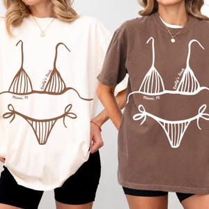 Pode incluir: Uma t-shirt creme com um gráfico de biquíni castanho e uma t-shirt castanha com um gráfico de biquíni branco. Ambas apresentam o texto Emily's Bach e Miami, Fl. Os designs de biquíni consistem num top triangular e cuecas com laços laterais.