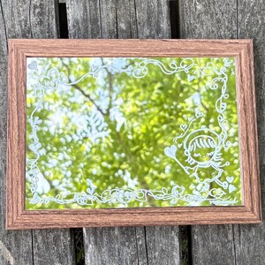 Könnte beinhalten: Ein rechteckiger Spiegel mit einem braunen Holzrahmen. Die Glasoberfläche ist mit weißen, handgemalten Linien verziert, die eine Bordüre aus geschwungenen Ranken und kleinen Herzen bilden, sowie einer zentralen Illustration einer Figur mit Hut, die ein Herz hält.