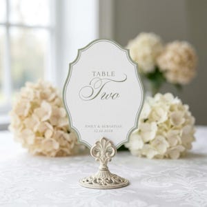 May include: A white scalloped table number card with a sage green border, featuring the text TABLE Two, the names EMILY & SEBASTIAN, and the date 12.12.2028, held upright by an ornate, distressed cream-colored metal stand with a decorative base.