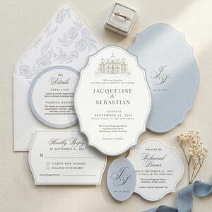 May include: A wedding invitation suite featuring a white envelope with a floral liner, a ring box, and several cards in white and light blue. The main invitation displays a line drawing of a manor, the names Jacqueline and Sebastian, and event details.