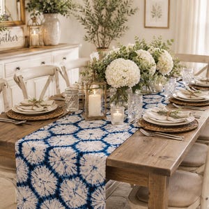 Puede incluir: Una larga mesa de comedor de madera preparada para una comida, con un camino de mesa tie-dye azul y blanco con patrón geométrico. La mesa está decorada con centros de mesa de hortensias blancas, farolillos de cristal y manteles individuales.