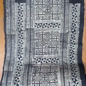May include: A black and white textile with a geometric pattern. The design features repeating cross-like shapes, triangles, and vertical stripes. The fabric appears to be woven, with a slightly rough texture. The overall aesthetic is a blend of traditional and modern design.