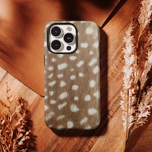 Puede incluir: Una funda rectangular para smartphone con un patrón de puntos marrones y blancos que imita la apariencia de la piel de ciervo. La funda tiene un recorte negro para la cámara con tres lentes circulares y un flash, con acabado liso.
