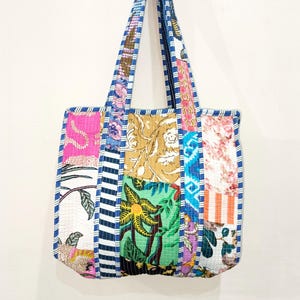 Op de afbeelding: Een patchwork tote bag met een verscheidenheid aan kleurrijke stoffen panelen. De tas heeft een mix van bloemen-, gestreepte en patroonontwerpen in roze, groen, blauw en bruin. De handvatten en randen zijn afgewerkt met blauw-wit gestreepte stof.