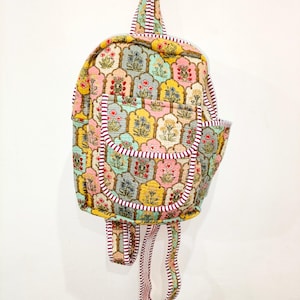 Puede incluir: Una mochila de tela acolchada con un patrón repetitivo de marcos arqueados que contienen motivos florales en amarillo, rosa, azul y verde. La bolsa incluye un bolsillo frontal, un asa superior y correas para los hombros con ribetes a rayas granates y blancas.